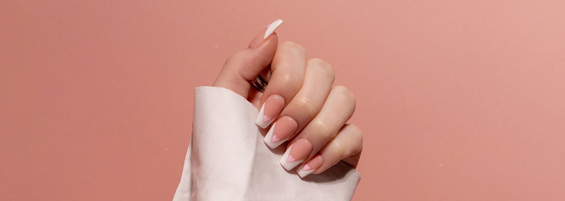 Nahaufnahme einer manikürten Hand mit French Nails und Herz-Nailart auf rosafarbenem Hintergrund – steht für sichere, TPO-freie Nagelpflege.